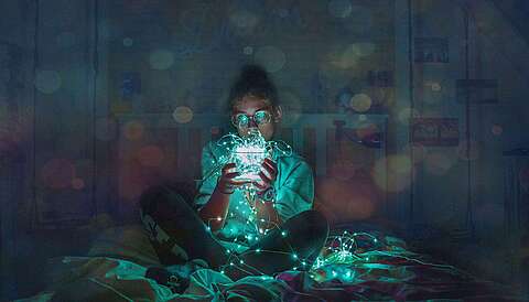 Auf einem Bett sitzendes Mädchen mit einem Glas in der Hand, in dem sich eine leuchtende Lichterkette befindet, die über das Glas hinausragt.
